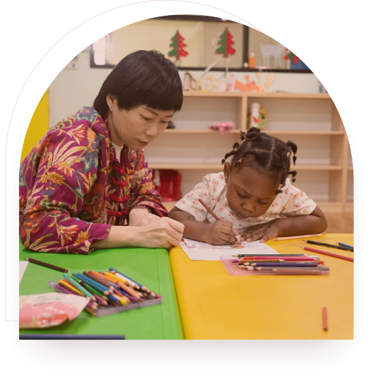 A clinician supporting a child colouring at a desk in a warm classroom setting