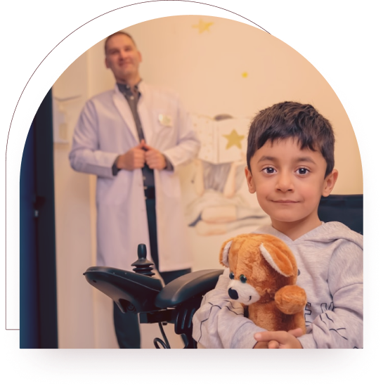 A young boy in a wheelchair holding a teddy bear, with a clinician standing supportively behind him