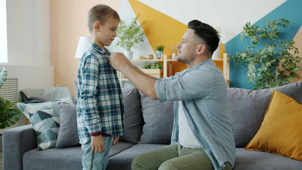 A father buttoning his son's shirt in a colourful living room