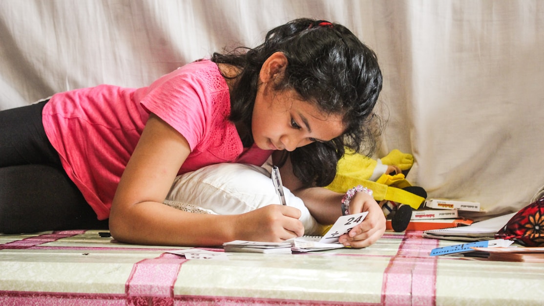 A girl lying down and writing in a notebook beside a soft toy