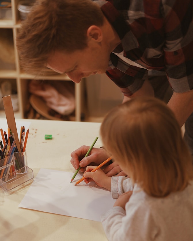 A man helping a young child draw at a table