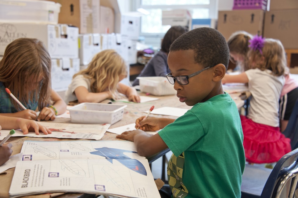 A boy wearing glasses drawing at a classroom desk