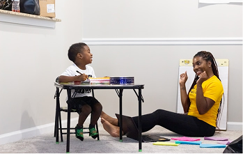 A boy sitting at a table with a clinician seated on the floor nearby