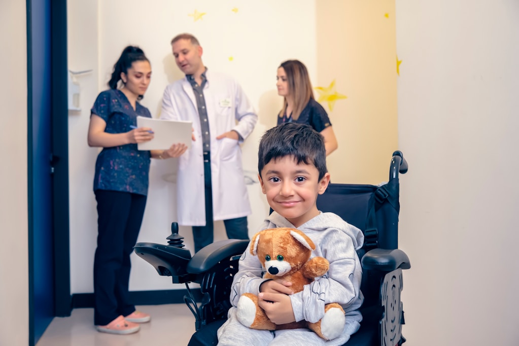 A boy in a wheelchair holding a teddy bear with clinical staff at Hiba by Anthalora