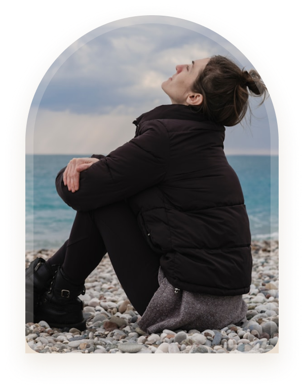 A young person sitting alone on a rocky beach hugging their knees