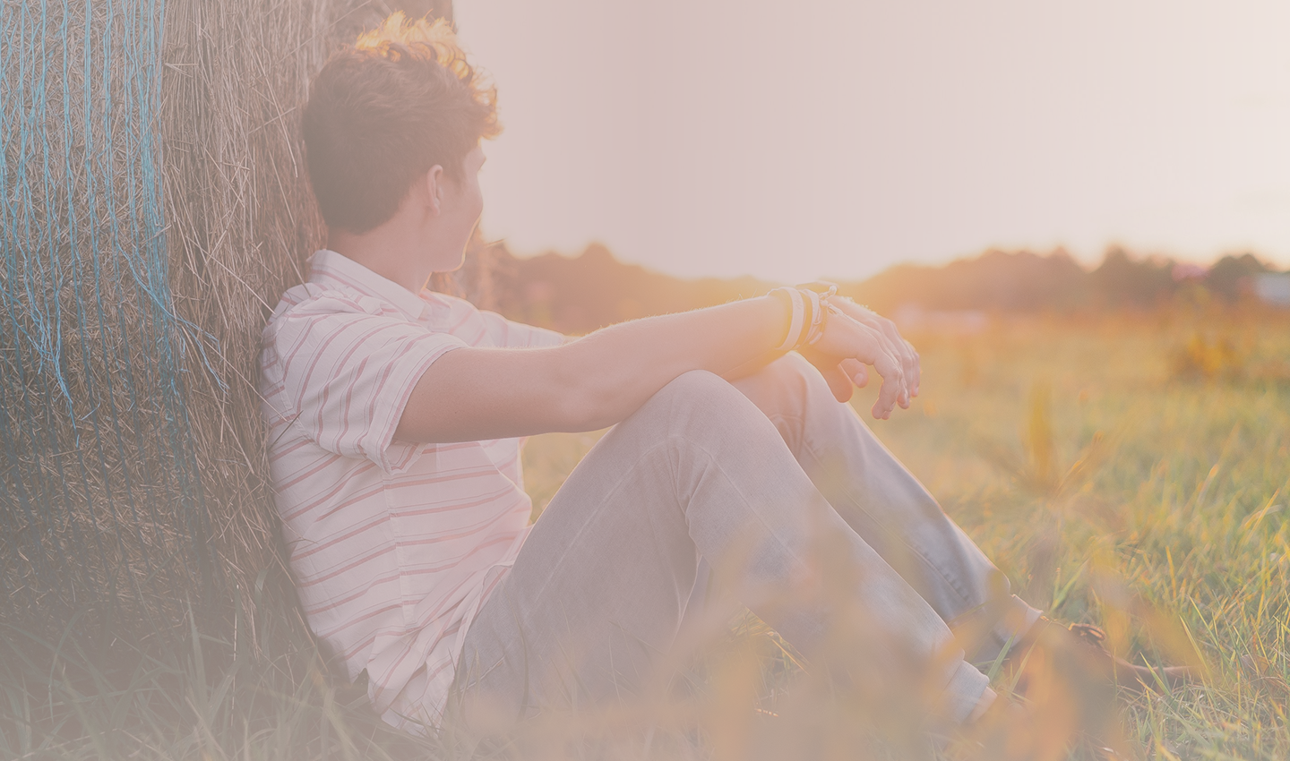 A young man sitting against a hay bale looking out across a field